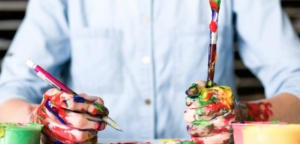 A man covered in paint holding a pencil in one hand and a paint brush in the other